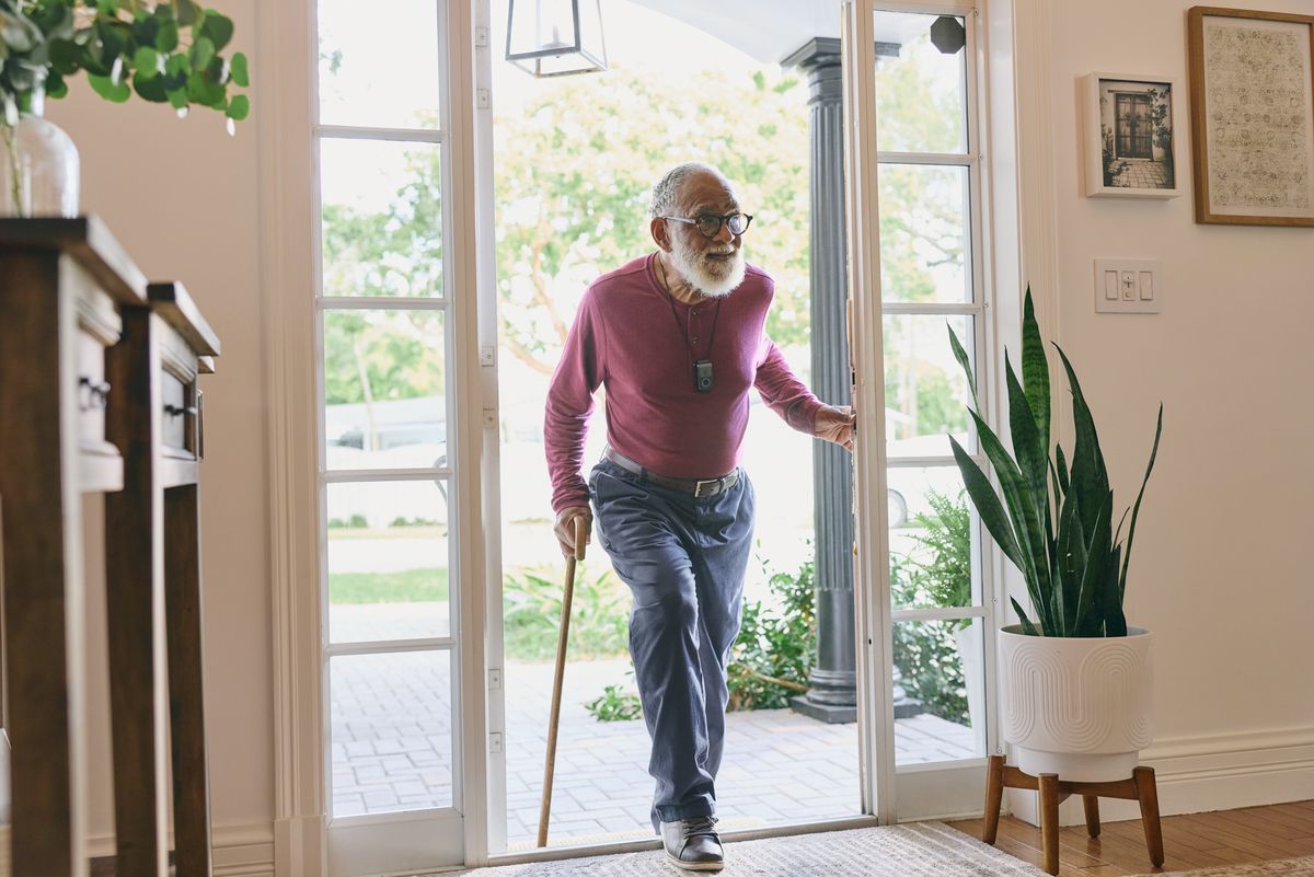 Older man with a cane wearing an Indigo pendant, stepping in through his front door